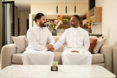 The intertwining of hands in a handshake and greeting between two men, spending enjoyable time with friends, discussion and dialogue among friends, youth entertainment and amusement, two Arab Gulf friends from Saudi Arabia wearing traditional thobes sitting on a comfortable white sofa, enjoying recreational activities on the weekend.