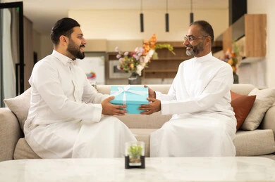 تبادل الهداية بين الأصدقاء في المناسبات والأعياد، تشابك أيدي مصافحة وسلام بين رجلين، قضاء وقت ممتع مع الأصحاب، المناقشة والحوار بين الأصدقاء، التسليه والترفيه الشبابي,  صديقان عربيان خليجيان سعوديان يرتديان الثوب التقليدي يجلسان على آريكه مريحة باللون الابيض
