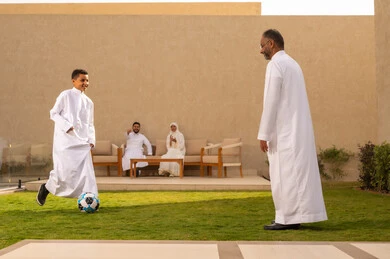 ايماءات طفل تدل على الحب و الفرح، قضاء أوقات عائلية ممتعة بالعطلة الصيفية في احدى الشاليهات, حب الآباء لأبنائهم، صورة من الخلف لجد عربي خليجي سعودي يرتدي الثوب الأبيض يلعب مع حفيده بالكرة, مفهوم الأسرة السعيدة، خلفية رجل وإمرأة يتبادلون الأحاديث