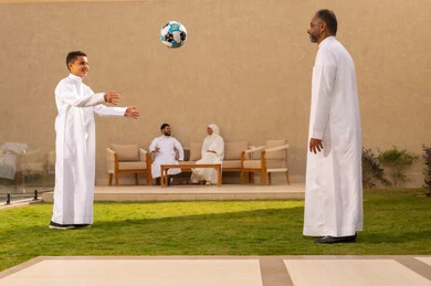 قضاء أوقات عائلية ممتعة بالعطلة الصيفية في احدى الشاليهات, ايماءات طفل تدل على الحب و الفرح،  حب الآباء لأبنائهم، صورة من الخلف لجد عربي خليجي سعودي يرتدي الثوب الأبيض يلعب مع حفيده بالكرة, مفهوم الأسرة السعيدة، خلفية رجل وإمرأة يتبادلون الأحاديث