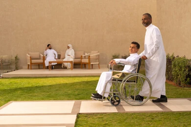 تحسين حياة أصحاب الهمم وتأمين احتياجاتهم الخاصة, أدوات تساعد مرضى ذوي الإعاقة الحركية على التنقل بسهولة, رجل مسن عربي خليجي سعودي يرتدي الزي التقليدي برفقة ابنه في إحدى الحدائق للاستمتاع بالهواء الطلق، ولد سعودي مقعد يجلس على كرسي متحرك، خلفية رجل وإمراة