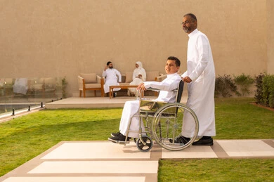 أدوات تساعد مرضى ذوي الإعاقة الحركية على التنقل بسهولة, تحسين حياة أصحاب الهمم وتأمين احتياجاتهم الخاصة, رجل مسن عربي خليجي سعودي يرتدي الزي التقليدي برفقة ابنه في إحدى الحدائق للاستمتاع بالهواء الطلق، ولد سعودي مقعد يجلس على كرسي متحرك يمد يديه جانبا
