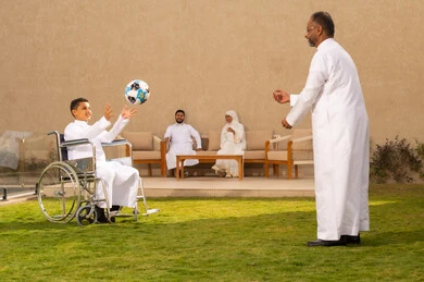 Playing with a Saudi boy in a wheelchair, improving the lives of people with disabilities and securing their special needs, tools that help patients with mobility disabilities to move easily, an elderly Arab Gulf Saudi man accompanied by his son in one of the chalets to play and enjoy the fresh air, background of a man and a woman.