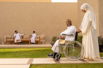 الاخلاص والعطف بين الزوجين ،جودة حياة ذوي الهمم في السعودية ، تأمين أدوات ومعدات لمساعدة المرضي ، رجل مقعد يجلس على كرسي متحرك، امرأة عربية خليجية سعودية ترتدي العباءة والحجاب تساعد زوجها على التنقل والحركة، خلفية اب مع طفله يتبادلون الأحاديث.