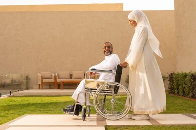 تبادل الأحاديث خلال التنزه، جودة حياة ذوي الهمم في السعودية ، الاخلاص والعطف بين الزوجين، تأمين أدوات ومعدات لمساعدة المرضي ، رجل مقعد يجلس على كرسي متحرك، امرأة عربية خليجية سعودية ترتدي العباءة والحجاب تساعد زوجها على التنقل والحركة.
