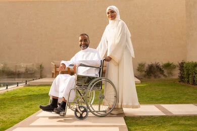 Looking at the camera with gestures of affection and love, the quality of life for people with disabilities in Saudi Arabia, loyalty and compassion between spouses, providing tools and equipment to assist patients, a disabled man sitting in a wheelchair, an Arab Gulf Saudi woman wearing an abaya and hijab helping her husband to move and navigate.