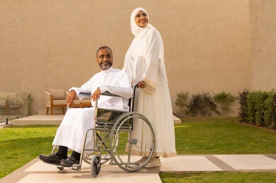 النظر إلى شيء ما في الأعلى، تأمين أدوات ومعدات لمساعدة المرضي، جودة حياة ذوي الهمم في السعودية ، الاخلاص والعطف بين الزوجين،  رجل مقعد يجلس على كرسي متحرك، امرأة عربية خليجية سعودية ترتدي العباءة والحجاب تساعد زوجها على التنقل والحركة.