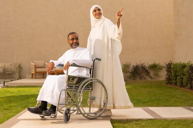 Referring to something above, securing tools and equipment to assist patients, the quality of life for people with disabilities in Saudi Arabia, loyalty and compassion between spouses, a disabled man sitting in a wheelchair, an Arab Gulf Saudi woman wearing an abaya and hijab helping her husband to move and navigate.
