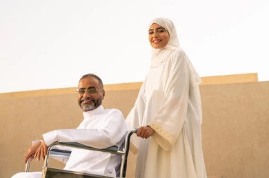 A smile and a look at the camera, securing tools and equipment to assist patients, the quality of life for people with disabilities in Saudi Arabia, loyalty and compassion between spouses, a disabled man sitting in a wheelchair, an Arab Gulf Saudi woman wearing an abaya and hijab helping her husband to move and navigate.