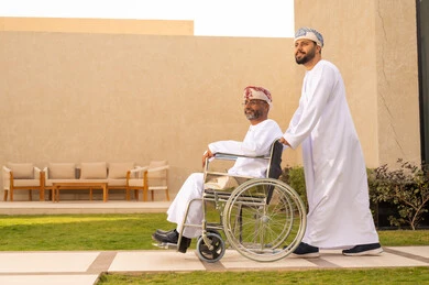 إيماءات السعادة والسرور بالخروج في الهواء الطلق ، أسلوب حياة أصحاب الهمم، تأمين أدوات ومعدات تساعد ذوى الهمم ، نمط حياة ذوي الإعاقة الحركية ، رجل مقعد يجلس على كرسي متحرك، شاب عربي خليجي عماني يرتدي الدشداشة والكمة ، يساعد والده على التنقل والحركة