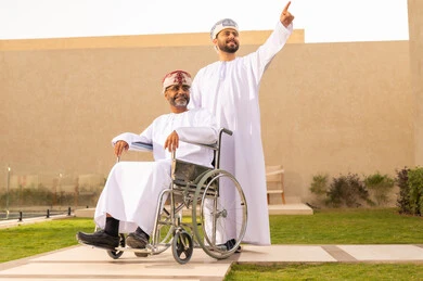 رجل يشير إلى شيء ما، تأمين أدوات ومعدات تساعد ذوى الهمم ، إيماءات السعادة والسرور بالخروج في الهواء الطلق ، أسلوب حياة أصحاب الهمم، نمط حياة ذوي الإعاقة الحركية ، رجل مقعد يجلس على كرسي متحرك، شاب عربي خليجي عماني يرتدي الدشداشة والكمة ، يساعد والده على التنقل