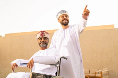التحدث والأشارة إلى شيء ما، تأمين أدوات ومعدات تساعد ذوى الهمم ، إيماءات السرور بالخروج في الهواء الطلق ، أسلوب حياة أصحاب الهمم، نمط حياة ذوي الإعاقة الحركية ، رجل مقعد يجلس على كرسي متحرك، شاب عربي خليجي عماني يرتدي الدشداشة والكمة ، يساعد والده على التنقل