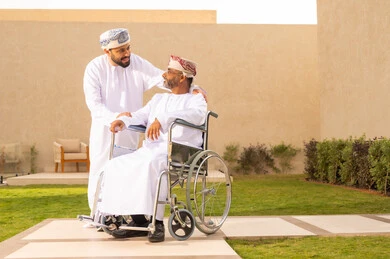 الاطمئنان في حال احتياج شيء، إيماءات السرور بالخروج في الهواء الطلق، تأمين أدوات ومعدات تساعد ذوى الهمم ، أسلوب حياة أصحاب الهمم، نمط حياة ذوي الإعاقة الحركية ، رجل مقعد يجلس على كرسي متحرك، شاب عربي خليجي عماني يرتدي الدشداشة والكمة ، يساعد والده على التنقل