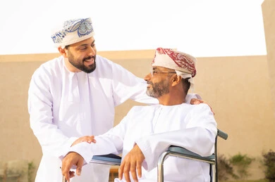 The question in case of needing something, gestures of joy when going outdoors, securing tools and equipment to assist people with disabilities, the lifestyle of people with disabilities, the lifestyle of individuals with mobility impairments, a seated man sitting in a wheelchair, a young Arab Gulf Omani man wearing a dishdasha and a kumma, helping his father to move around.