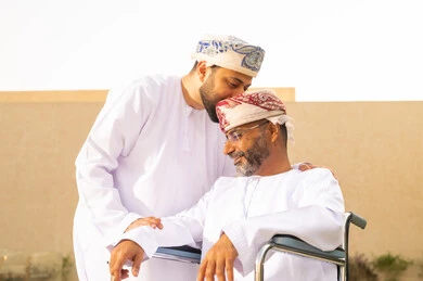 قبلة رأس من ولر لوالده، إيماءات السرور بالخروج في الهواء الطلق، تأمين أدوات ومعدات تساعد ذوى الهمم ، أسلوب حياة أصحاب الهمم، نمط حياة ذوي الإعاقة الحركية ، رجل مقعد يجلس على كرسي متحرك، شاب عربي خليجي عماني يرتدي الدشداشة والكمة ، يساعد والده على التنقل