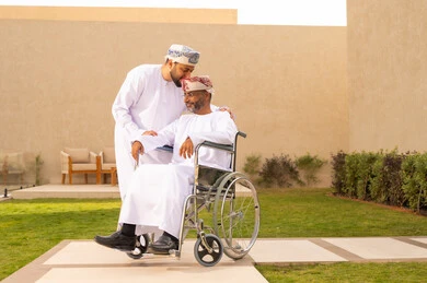 A kiss on the head is a sign of respect and appreciation, gestures of joy when going outdoors, securing tools and equipment to assist people with disabilities, the lifestyle of people with disabilities, the lifestyle of individuals with mobility impairments, a seated man sitting in a wheelchair, a young Arab Gulf Omani man wearing a dishdasha and kumma, helping his father to move around.