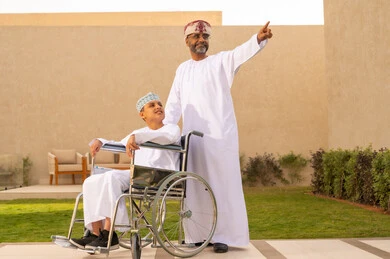 الأشارة إلى شيء ما ، عائلة عربية عمانية خليجية في رحلة بإحدى الشاليهات في سلطنه عمان، مشاركة الأبناء لحظات ممتعة في العطلة، صورة مقربة لولد عربي خليجي عماني يرتدي الدشداشة و الكمه يجلس على كرسي متحرك، رجل عربي عماني يساعد ابنه على التنقل والحركة