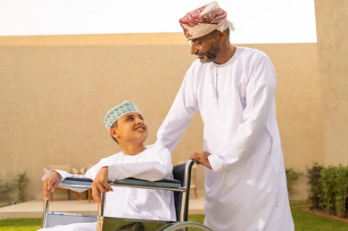 Spending time during the summer holidays, an Arab Omani Gulf family on a trip to one of the chalets in the Sultanate of Oman, sharing enjoyable moments with the children during the vacation. A close-up image of an Arab Gulf Omani boy wearing a dishdasha and a kumma sitting in a wheelchair, an Arab Omani man helping his son to move and navigate.