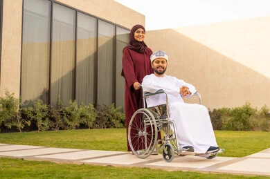 الاخلاص والعطف بين الزوجين ،جودة حياة ذوي الهمم في السعودية ، تأمين أدوات ومعدات لمساعدة المرضي ، رجل مقعد يجلس على كرسي متحرك، امرأة عربية خليجية سعودية ترتدي العباءة والحجاب تساعد زوجها على التنقل والحركة ،  قضاء الوقت في المنتزه مع العائلة