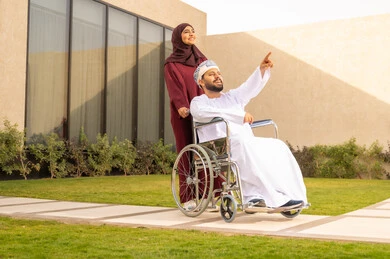 The quality of life for people with disabilities in Saudi Arabia, the affection and compassion between spouses, providing tools and equipment to assist patients, a disabled man sitting in a wheelchair, an Arab Gulf Saudi woman wearing an abaya and hijab, helping her husband to move and navigate, spending time in the park with family, nature background.