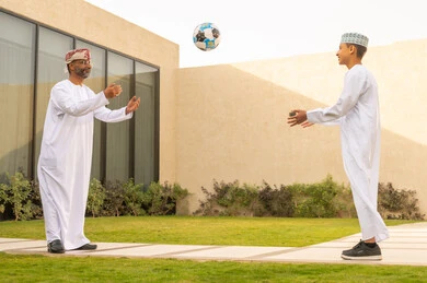 Gestures of a child indicating love and joy, spending enjoyable family time during the summer vacation in one of the chalets, the love of parents for their children, a picture from behind of an Arab Gulf Omani grandfather wearing a white robe playing with his grandson with a ball, the concept of a happy family, a green nature background in one of the Omani chalets.