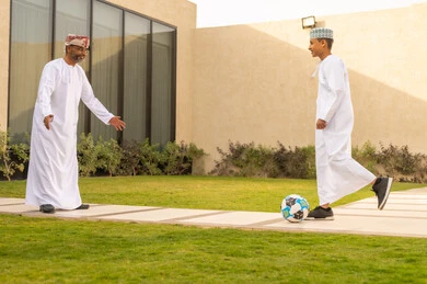A green nature background in one of the Omani chalets, gestures of a child indicating love and joy, spending enjoyable family time during the summer vacation in one of the chalets, the love of parents for their children, a picture from behind of an Arab Gulf Omani grandfather wearing a white dishdasha playing with his grandson with a ball, the concept of a happy family.