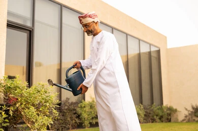 استخدام مرش المياة ، العناية بالنباتات فى الحديقة ، رجل عربي خليجي عماني يرتدي الدشداشة والكمة يقوم باستخدام وعاء السقي في ري النباتات ، اجواء الطبيعة الخلابة ، مفهوم النظافة والاهتمام بحديقة المنزل تهدئة الأعصاب و النفسية من خلال أنشطة مهارات الحياة.
