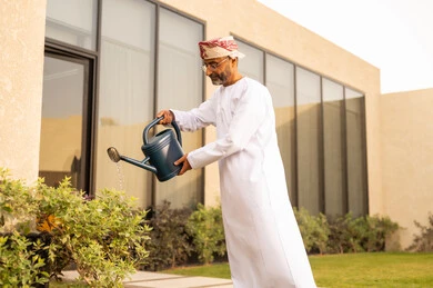 العناية بالنباتات فى الحديقة ، استخدام مرش المياة ،  رجل عربي خليجي عمانييرتدب الدشداشة والكمة يقوم باستخدام وعاء السقي في ري النباتات ، اجواء الطبيعة الخلابة ، مفهوم النظافة والاهتمام بحديقة المنزل تهدئة الأعصاب و النفسية من خلال أنشطة مهارات الحياة.