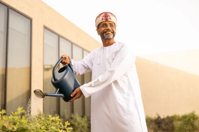 نظرات التأمل والنقاء، العناية بالنباتات فى الحديقة ، استخدام مرش المياه، رجل عربي خليجي عماني يرتدي الدشداشة والعمامة يقوم باستخدام وعاء السقي في ري النباتات، مفهوم النظافة والاهتمام بحديقة المنزل.