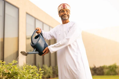 Standing outdoors, calming the nerves through life skills activities, taking care of the plants in the garden, using a watering can, an Arab Gulf Omani man using a watering pot to water the plants, the beautiful natural atmosphere, the concept of cleanliness and caring for the home garden.