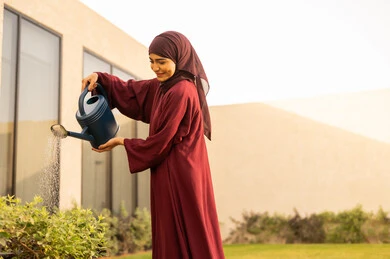 The concept of cleanliness and caring for the home garden, watering plants by spraying, a Saudi Gulf Arab woman using a watering can, an Omani Gulf Arab woman looking at the plants with gestures of happiness, the beautiful natural atmosphere, spending enjoyable time in one of the chalets, fun, entertainment, and recreation.