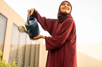 Looking up with gestures of happiness, the concept of cleanliness and caring for the home garden, a Saudi Gulf Arab woman using a watering can, an Omani Gulf Arab woman watering plants with a spray, the beautiful natural atmosphere, spending enjoyable time in one of the chalets, fun and entertainment.