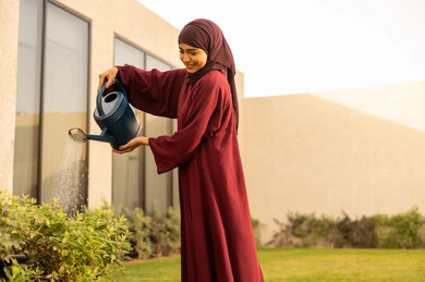 Watering plants by spraying, a Saudi Arabian Gulf woman using a watering can, the concept of cleanliness and caring for the home garden, an Omani Gulf woman looking at something with gestures of happiness, the beautiful natural atmosphere, spending enjoyable time in one of the chalets, fun, entertainment, and recreation.