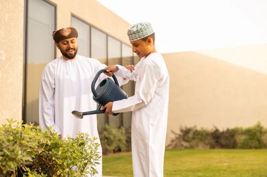 فعاليات وأجواء ترفيهية ، ايماءات السعادة والسرور، المشاركة مع الاولاد في المهم اليومية، قضاء الوقت الممتع في إحدى الشاليهات ، مفهوم النظافة والاهتمام بحديقة المنزل ، رجل عربي خليجي عماني يقضي وقت ممتع مع إبنه في ري النباتات بإستخدام وعاء السقي .