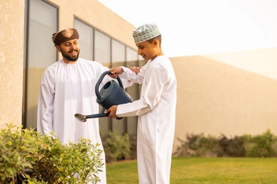 The concept of cleanliness and caring for the home garden, activities and recreational atmosphere, expressions of happiness on their faces, participating with the children in daily tasks, spending enjoyable time in one of the chalets, an Arab Gulf Omani man enjoying time with his son watering the plants using a watering can.