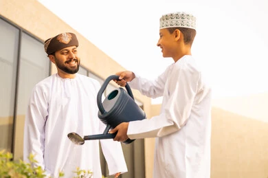 Participating with the children in daily tasks, spending enjoyable time in one of the chalets, the concept of cleanliness and caring for the home garden, an Arab Gulf Omani man wearing a dishdasha and turban enjoying time with his son watering the plants using a watering can, gestures of happiness and joy.