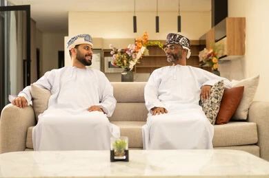 Chatting and exchanging conversations, enjoying spending time with children, an elderly Arab Gulf Omani man wearing a dishdasha and turban sitting on the sofa next to his young son in the living room, happy family gatherings, expressions of affection and love.