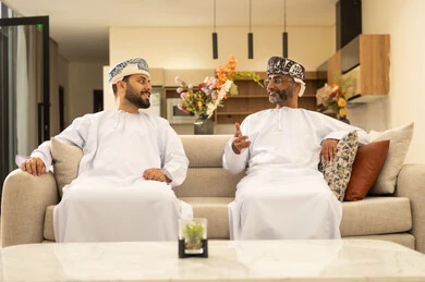 Enjoying time with the children, an elderly Arab Gulf Omani man wearing a dishdasha and turban sits on the sofa next to his young son in the living room, sharing stories and conversations, happy family gatherings, expressions of affection and love.