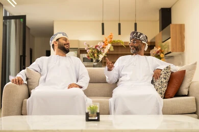 جلسات عائلية سعيدة، تعابير المودة والمحبة، التسامر وتبادل الاحاديث، الاستمتاع بقضاء وقت مع الابناء، رجل كبير بالسن عربي خليجي عماني يرتدي الدشداشة والعمامة يجلس على الاريكة بجانب ابنه الشاب في غرفة الجلوس