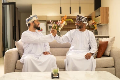 Chatting and exchanging conversations, enjoying spending time with children, an elderly Arab Gulf Omani man wearing a dishdasha and turban sitting on the sofa next to his young son in the living room, happy family gatherings, expressions of affection and love.