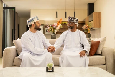 Chatting and exchanging conversations, enjoying spending time with children, an elderly Arab Gulf Omani man wearing a dishdasha and turban sitting on the sofa next to his young son in the living room, happy family gatherings, expressions of affection and love.