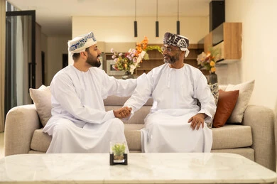 Enjoying time with the children, an elderly Arab Gulf Omani man wearing a dishdasha and turban sits on the sofa next to his young son in the living room, sharing stories and conversations, happy family gatherings, expressions of affection and love.