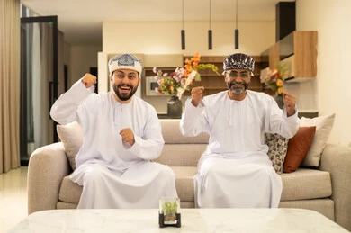 Interaction and engagement while watching the favorite team, matches, and sports games. Two Arab Omani Gulf men wearing the dishdasha and kumma, one of them standing with gestures of excitement, victory, and triumph, a passion for recreational activities.