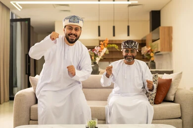 التفاعل والاندماج بمشاهدة الفريق المفضل, المباريات والالعاب الرياضية, رجلان عربيان عمانيان خليجيان يرتديان الدشداشة والكمة احدهما يقف بإيماءات الحماس و الفوز والانتصار، الشغف بالانشطة الترفيهية