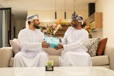Giving gifts on happy occasions, earning the parents' approval, happy family gatherings, expressions of affection and love, enjoying time spent with children, an elderly Arab Gulf Omani man wearing a dishdasha and turban sitting on the sofa next to his young son in the living room, a young man presenting a gift to his father.