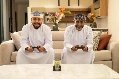 Gestures of happiness and excitement, participation and competition in modern technological games, two Arab Gulf men from Oman wearing the dishdasha and turban sitting on the sofa in the living room holding the game controller, the digital entertainment world, fun and entertainment with video games in the company of friends.