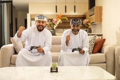 المشاركة والمنافسة بالألعاب التقنية الحديثة,  رجلان عربيان خليجيان عمانيان يرتديان الدشداشة والعمامة يجلسان على الاريكة في غرفة المعيشة يمسكان عصا التحكم, العالم الرقمي الترفيهي, المتعة والتسلية بالفيديو جيمز برفقة الأصدقاء، رفع قبضة اليد بايماءات السعادة والحماس