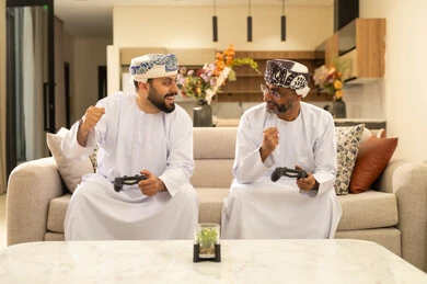 Gestures of happiness and excitement, participation and competition in modern technological games, two Arab Gulf men from Oman wearing the dishdasha and turban sitting on the sofa in the living room holding the game controller, the digital entertainment world, fun and entertainment with video games in the company of friends.