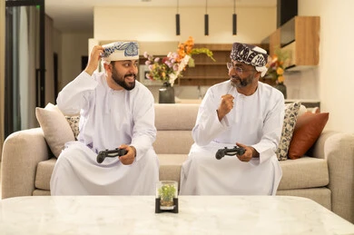 Raising a fist with gestures of happiness and enthusiasm, participating and competing in modern technological games, two Arab Gulf men from Oman wearing the dishdasha and turban sitting on the couch in the living room holding the game controller, the digital entertainment world, fun and entertainment with video games in the company of friends.