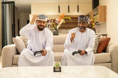 رفع قبضة اليد بايماءات السعادة والحماس، المشاركة والمنافسة بالألعاب التقنية الحديثة,  رجلان عربيان خليجيان عمانيان يرتديان الدشداشة والعمامة يجلسان على الاريكة في غرفة المعيشة يمسكان عصا التحكم, العالم الرقمي الترفيهي, المتعة والتسلية بالفيديو جيمز برفقة الأصدقاء